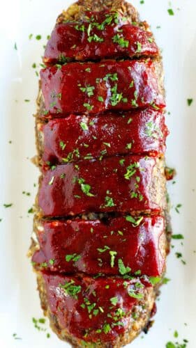liced meatloaf topped with a glossy, deep red glaze and sprinkled with fresh parsley on a white serving platter.