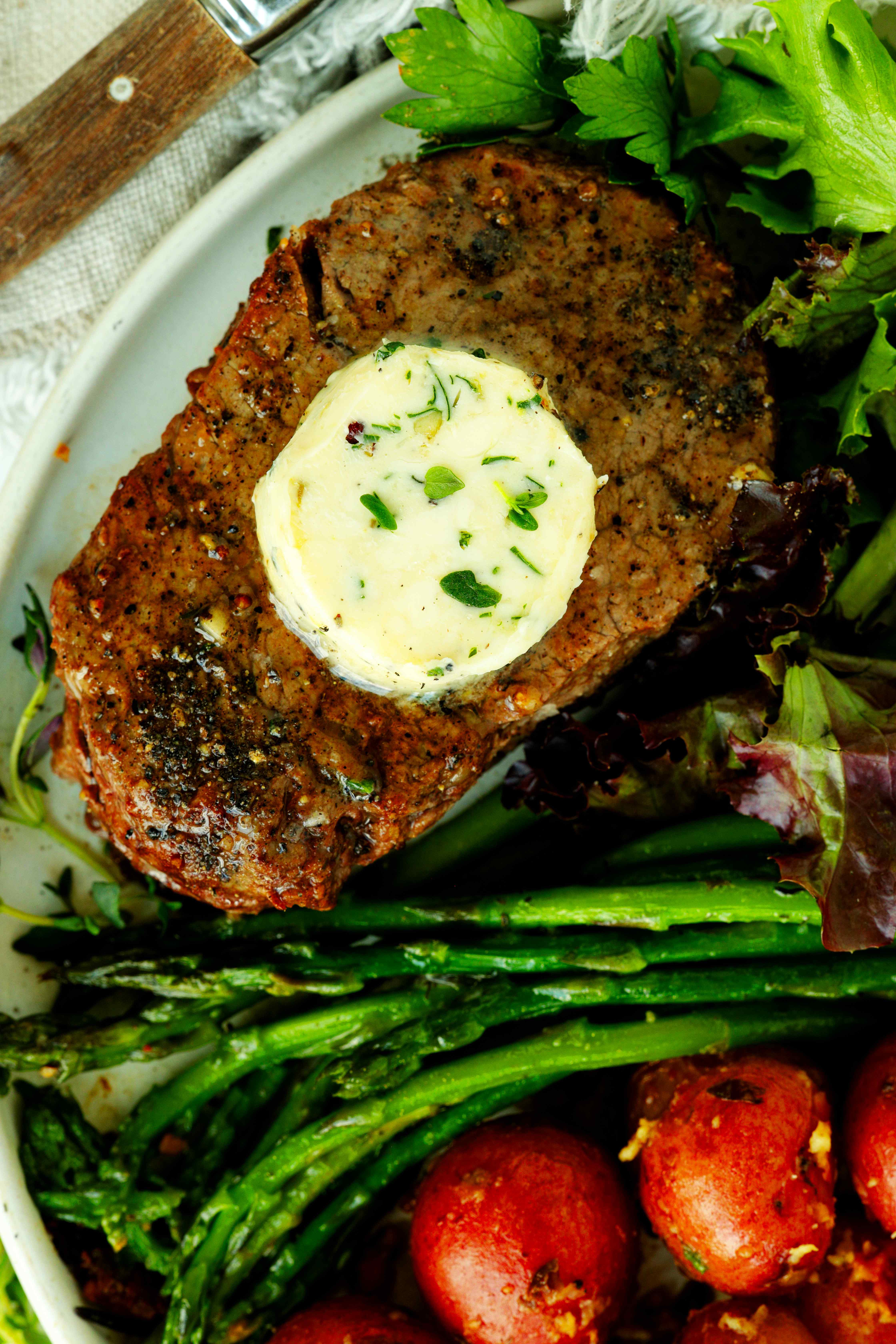 Juicy filet mignon steak topped with a round of herb butter, served with roasted baby red potatoes, asparagus, and fresh greens on a white plate.