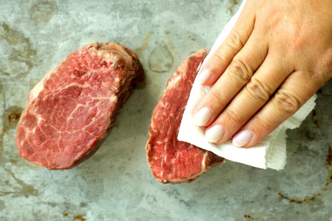 Hand using a paper towel to pat dry a raw filet mignon steak before cooking.