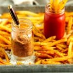 Seasoned French fries on a baking sheet with a jar of homemade fry seasoning and a jar of ketchup for dipping.