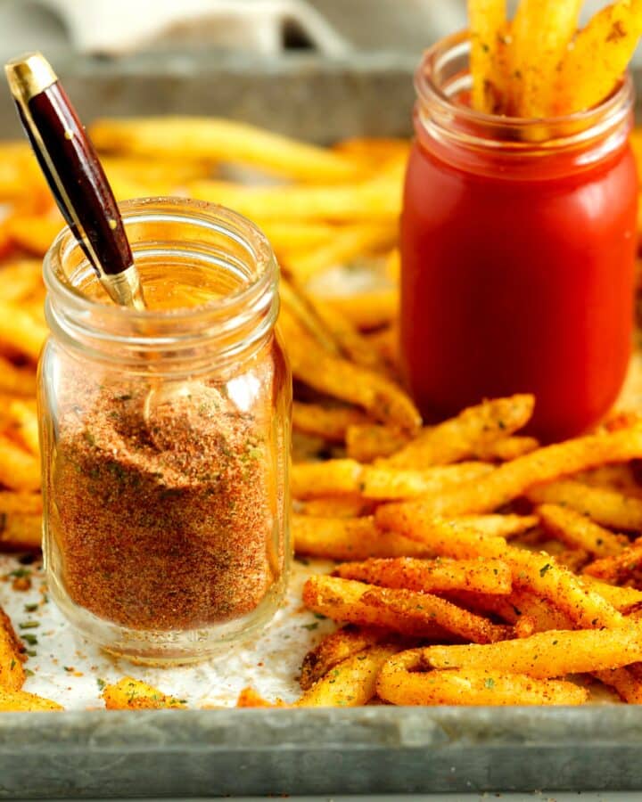 Seasoned French fries on a baking sheet with a jar of homemade fry seasoning and a jar of ketchup for dipping.