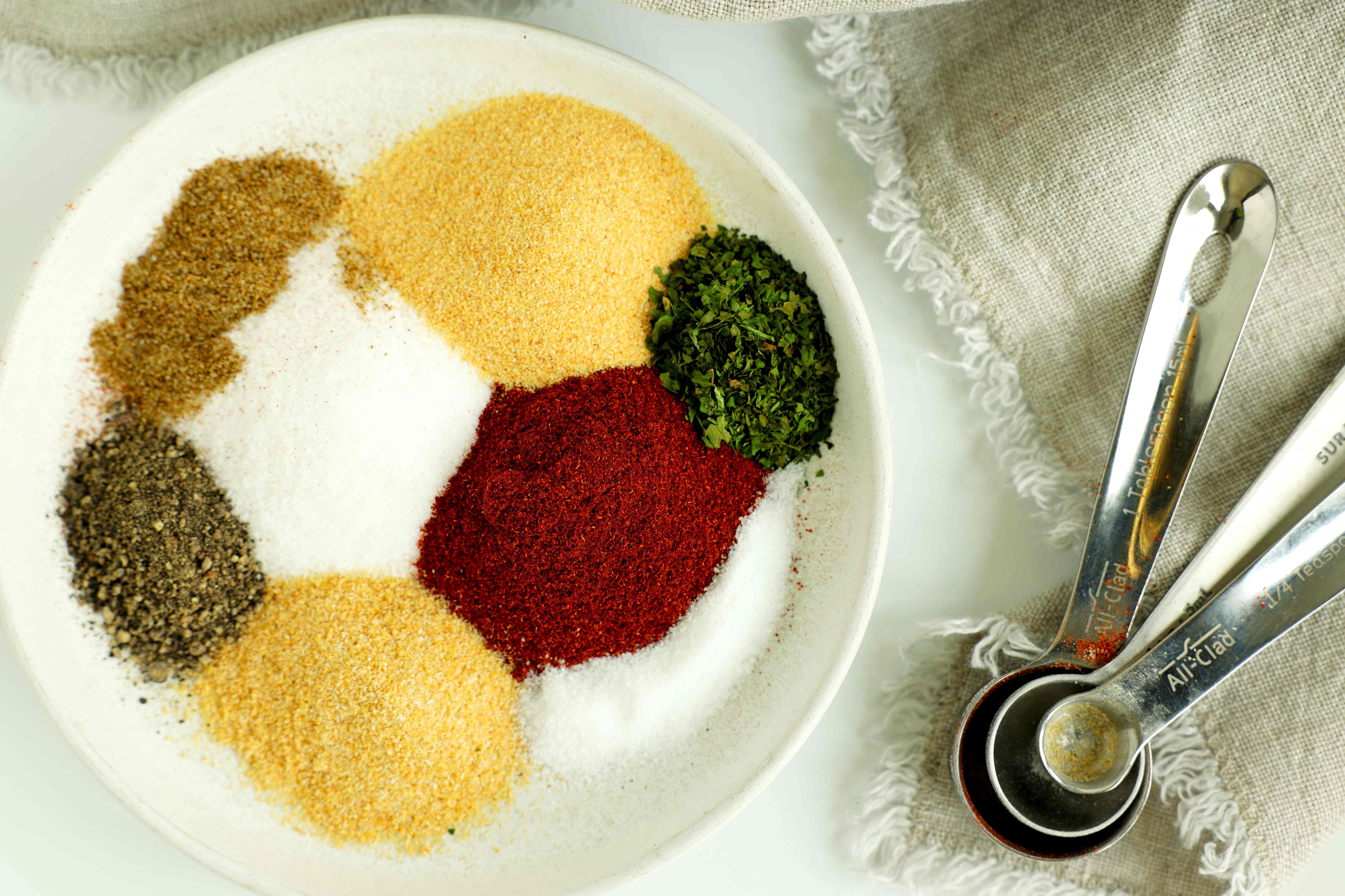 Assorted spices for homemade fry seasoning arranged in a white bowl, including paprika, garlic powder, onion powder, parsley, salt, and black pepper, with measuring spoons on the side.