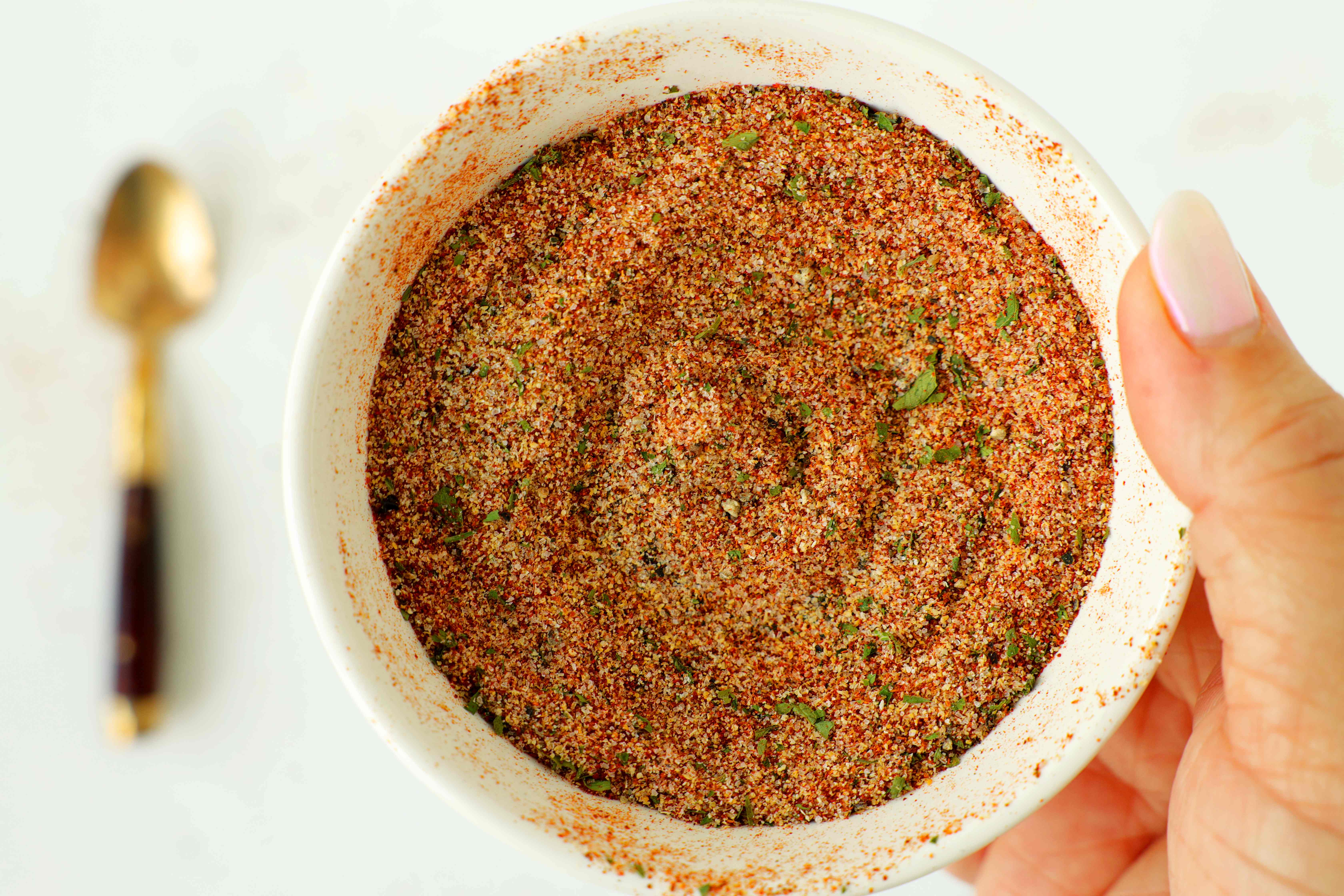 Homemade fry seasoning blend in a white bowl, showing a colorful mix of paprika, parsley, garlic, and spices, held in hand with a spoon blurred in the background.