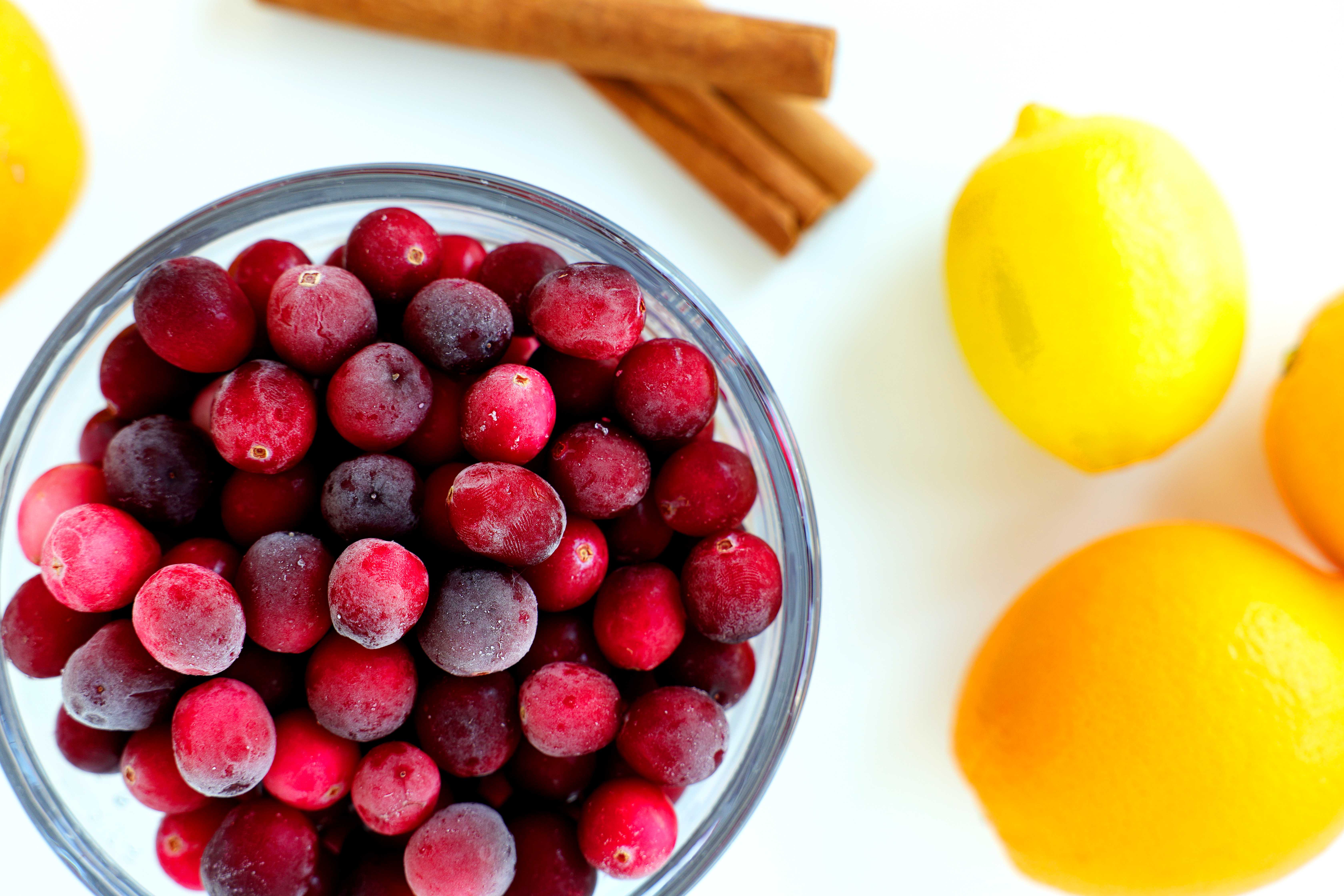 Fresh cranberries in a glass bowl surrounded by oranges, lemon, and cinnamon sticks — ingredients for homemade cranberry sauce with orange juice.