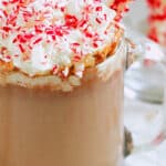 Close-up of peppermint hot chocolate in a clear mug topped with whipped cream and crushed candy canes, with a candy-cane stir stick.