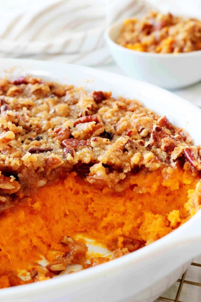 Sweet potato soufflé in a white casserole dish topped with a buttery pecan streusel; a large scoop removed reveals the bright orange, fluffy interior. A small bowl of the casserole sits blurred in the background.