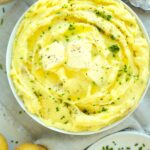 Wide overhead shot of mashed Yukon Gold potatoes in a white bowl on a neutral linen cloth, surrounded by herbs and potatoes.
