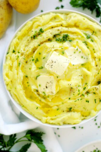 Top-down view of a serving bowl filled with creamy Yukon Gold mashed potatoes, garnished with butter, chives, and pepper on a linen background.
