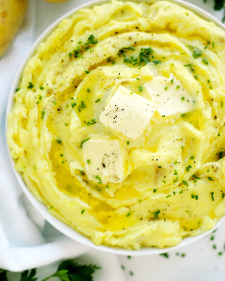 Top-down view of a serving bowl filled with creamy Yukon Gold mashed potatoes, garnished with butter, chives, and pepper on a linen background.