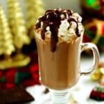 Festive dark hot chocolate in a clear footed mug with whipped cream, marshmallows, and chocolate sauce, surrounded by ornaments and chocolate pieces.