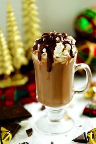 Festive dark hot chocolate in a clear footed mug with whipped cream, marshmallows, and chocolate sauce, surrounded by ornaments and chocolate pieces.