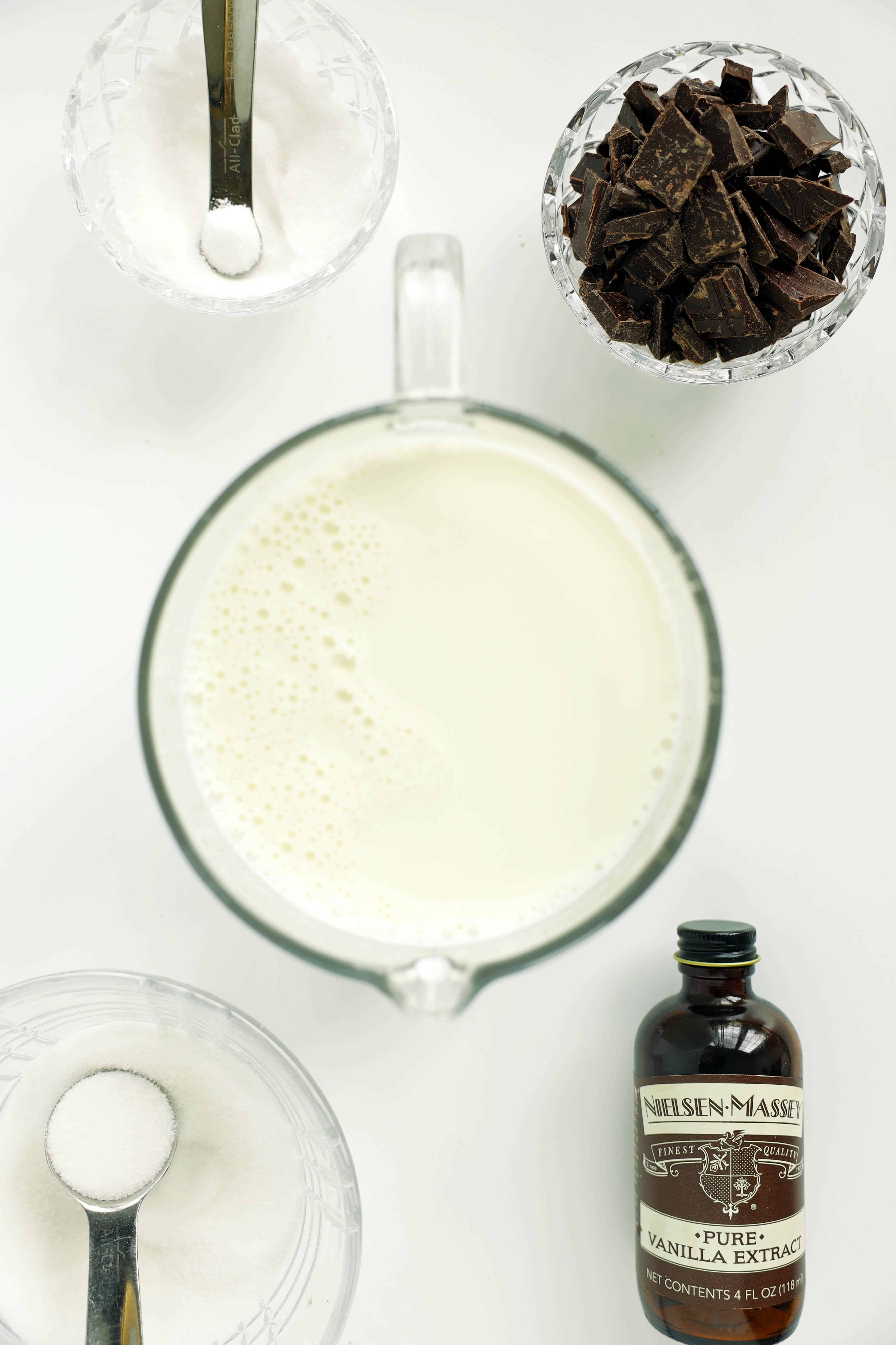 Overhead of dark hot chocolate ingredients&mdash;milk in a measuring jug, chopped chocolate, sugar and salt in spoons, and a bottle of vanilla.