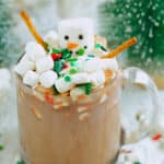 Glass mug of hot chocolate topped with a marshmallow snowman, mini marshmallows, sprinkles, and pretzel-stick arms; snowy bottle-brush trees blurred behind.