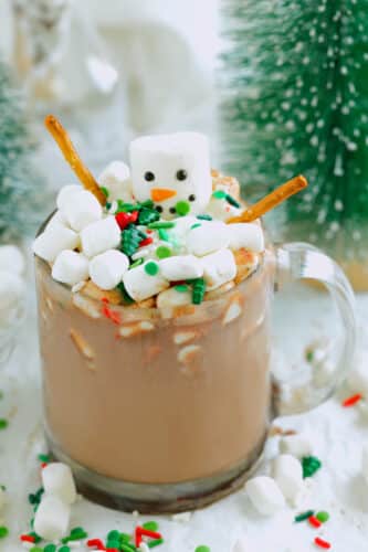 Glass mug of hot chocolate topped with a marshmallow snowman, mini marshmallows, sprinkles, and pretzel-stick arms; snowy bottle-brush trees blurred behind.