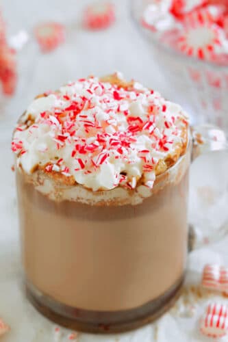 Peppermint hot chocolate in a clear mug topped with whipped cream and crushed candy canes, with peppermint candies blurred in the background.