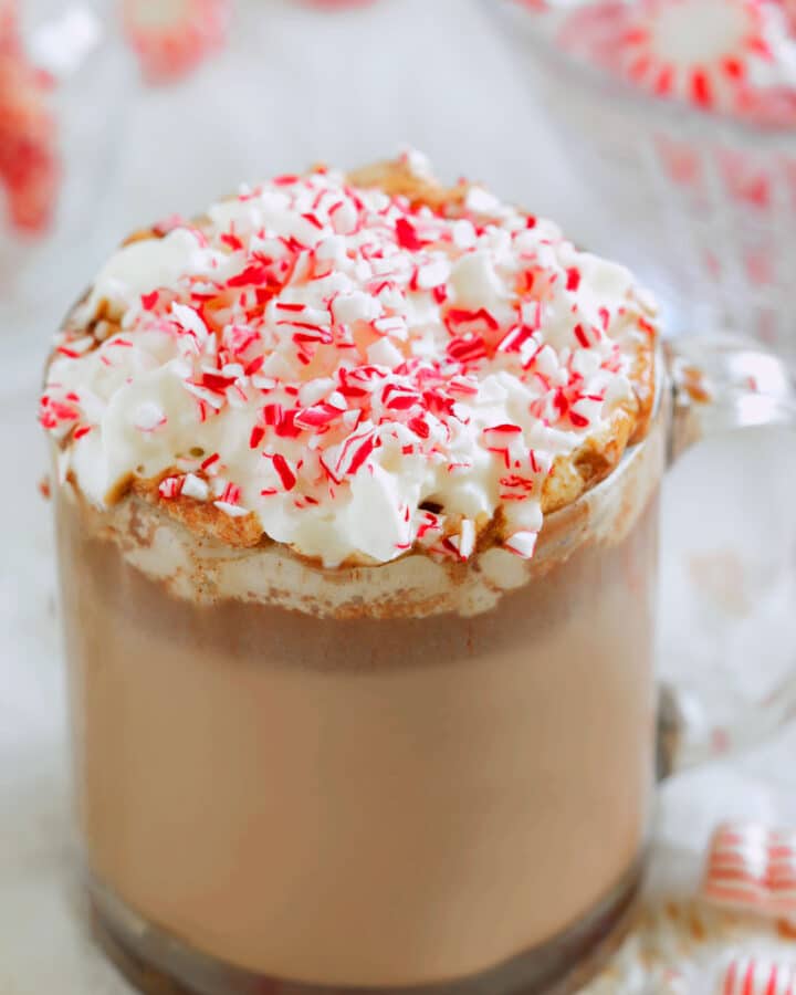 Peppermint hot chocolate in a clear mug topped with whipped cream and crushed candy canes, with peppermint candies blurred in the background.