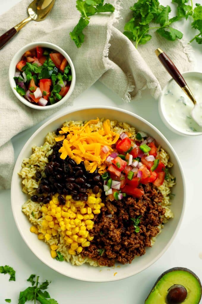 Taco bowl assembly with cilantro lime rice, taco meat, pico de gallo, cheese, corn, and beans.