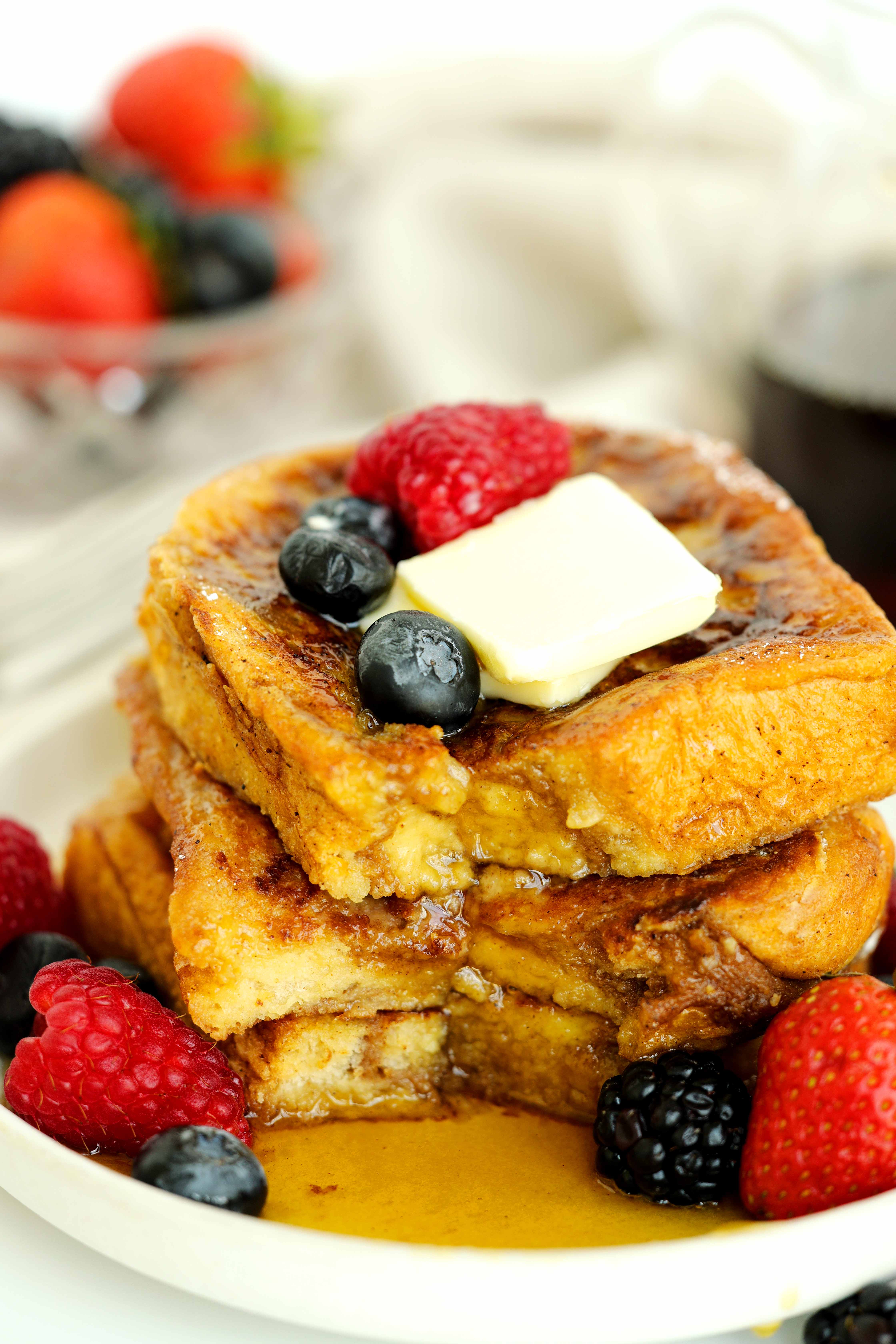 Stack of golden brioche French toast topped with butter, maple syrup, raspberries, blueberries, and blackberries on a white plate.