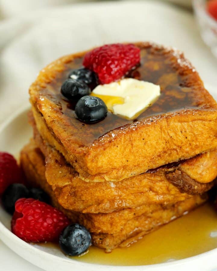 Stack of thick brioche French toast slices topped with butter, maple syrup, raspberries, and blueberries on a white plate, with fresh berries scattered around.