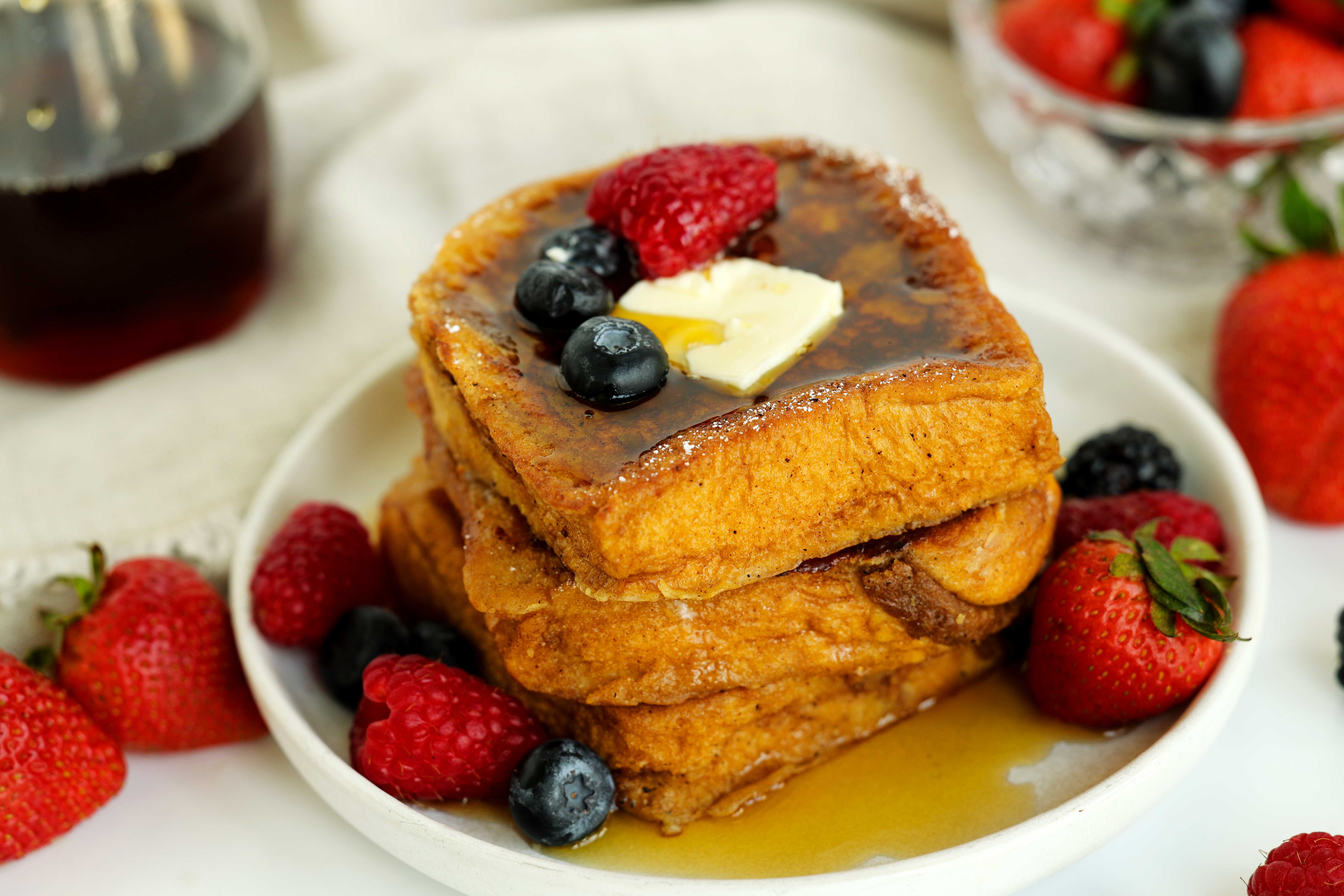 Stack of thick brioche French toast slices topped with butter, maple syrup, raspberries, and blueberries on a white plate, with fresh berries scattered around.