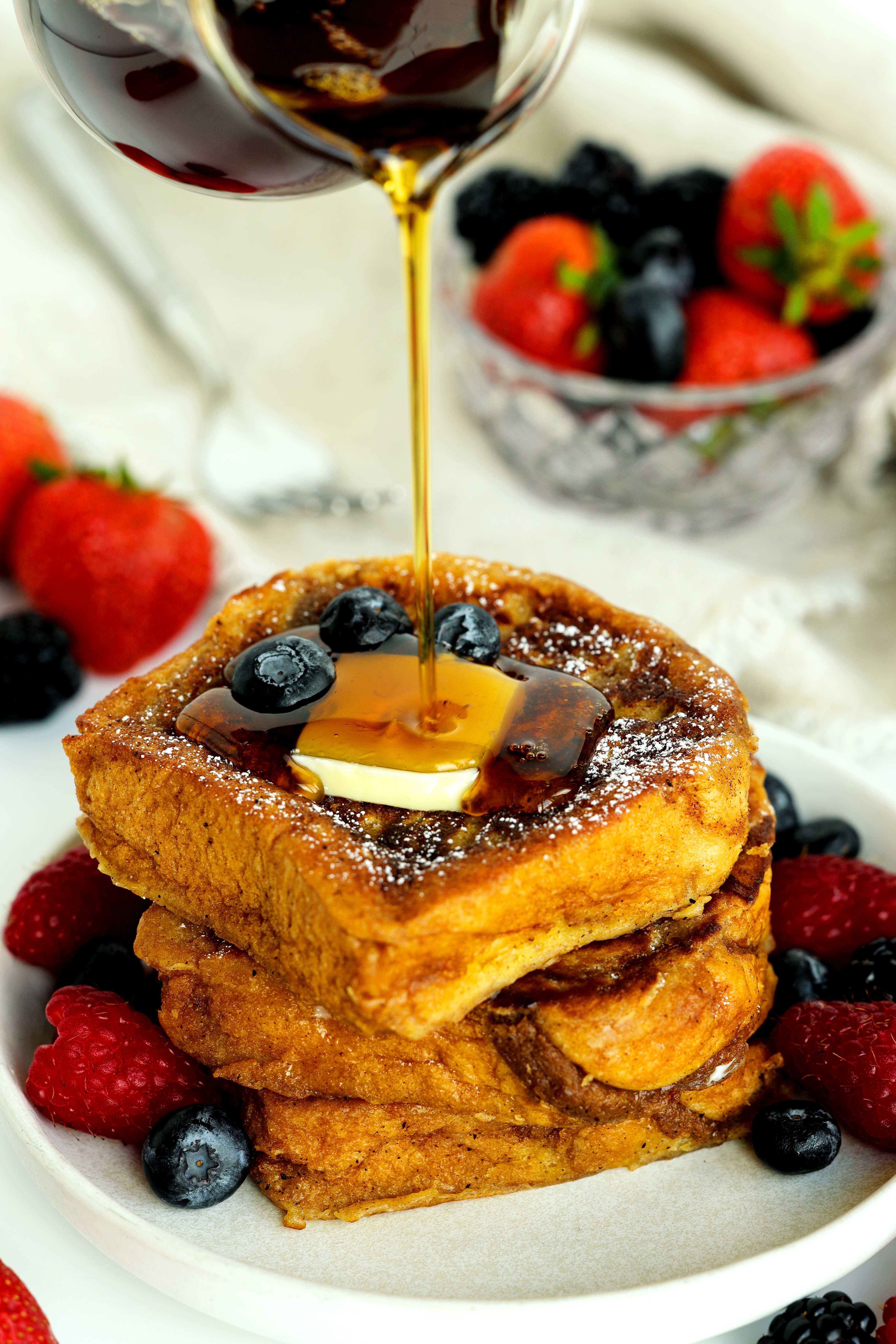 Maple syrup being poured over a stack of brioche French toast topped with butter and fresh berries on a white plate.
