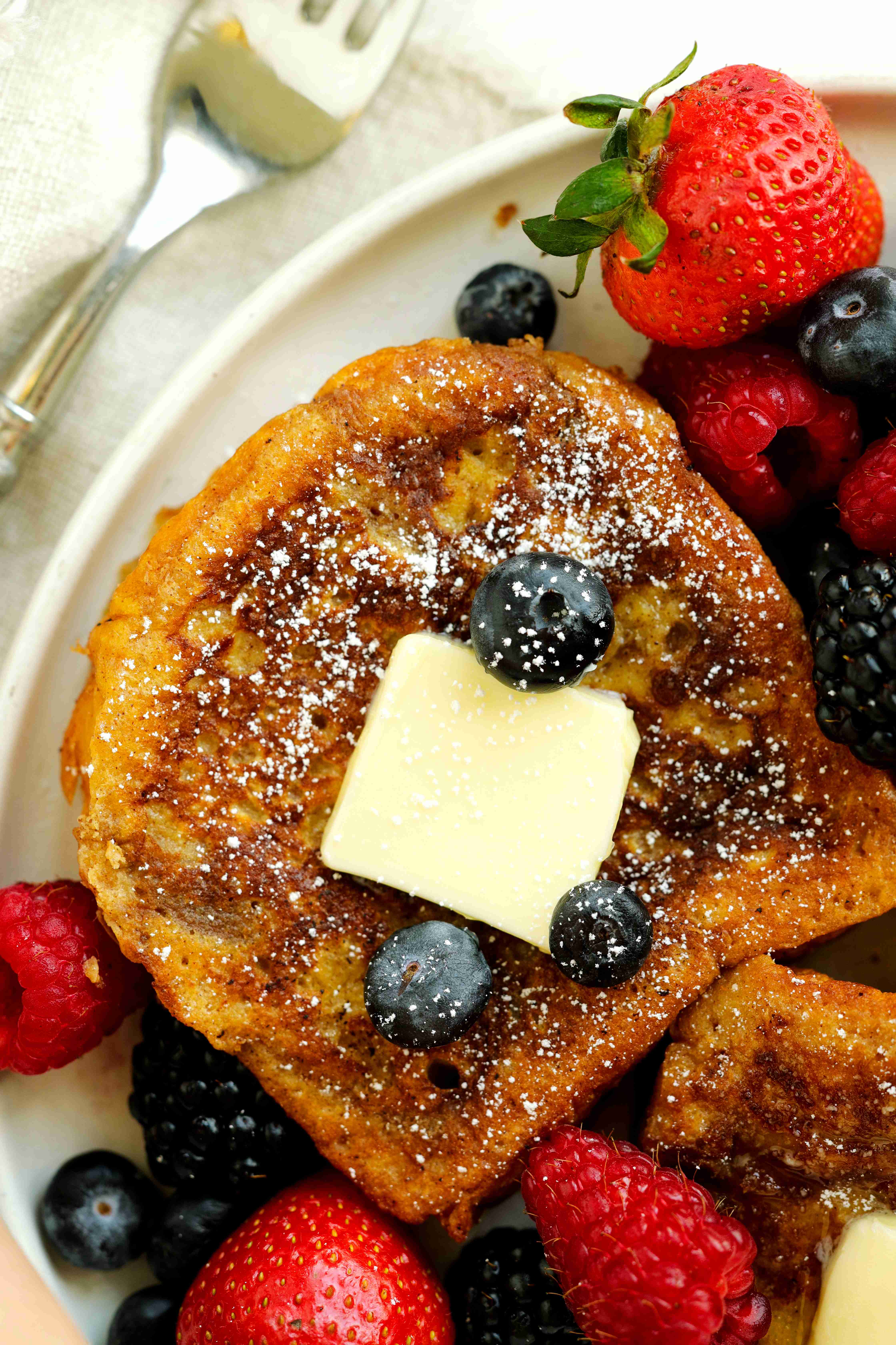 Golden brioche French toast topped with butter, powdered sugar, and fresh berries on a white plate.