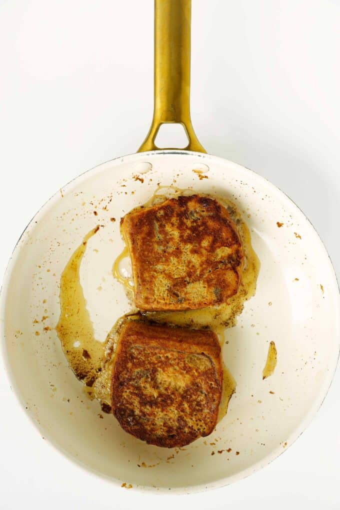 Two slices of brioche French toast cooking in butter in a skillet, showing deep golden-brown surfaces.