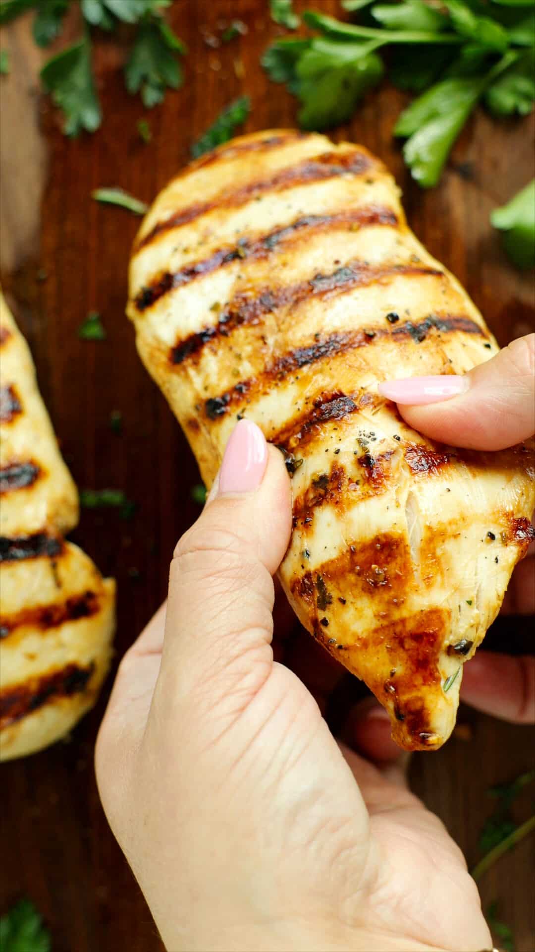 Grilled chicken breast with visible grill marks being pulled apart to show juicy interior, on a wooden board with fresh herbs.