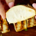 Close-up of sliced grilled chicken breast showing a juicy, fully cooked interior, held between fingers over a wooden board with additional sliced pieces in the background.