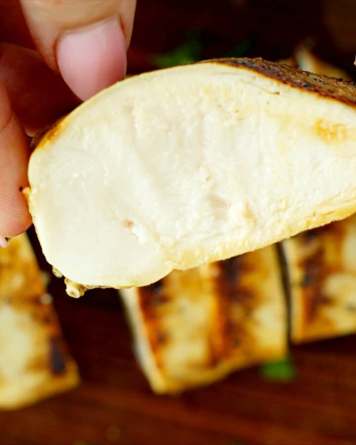 Close-up of sliced grilled chicken breast showing a juicy, fully cooked interior, held between fingers over a wooden board with additional sliced pieces in the background.