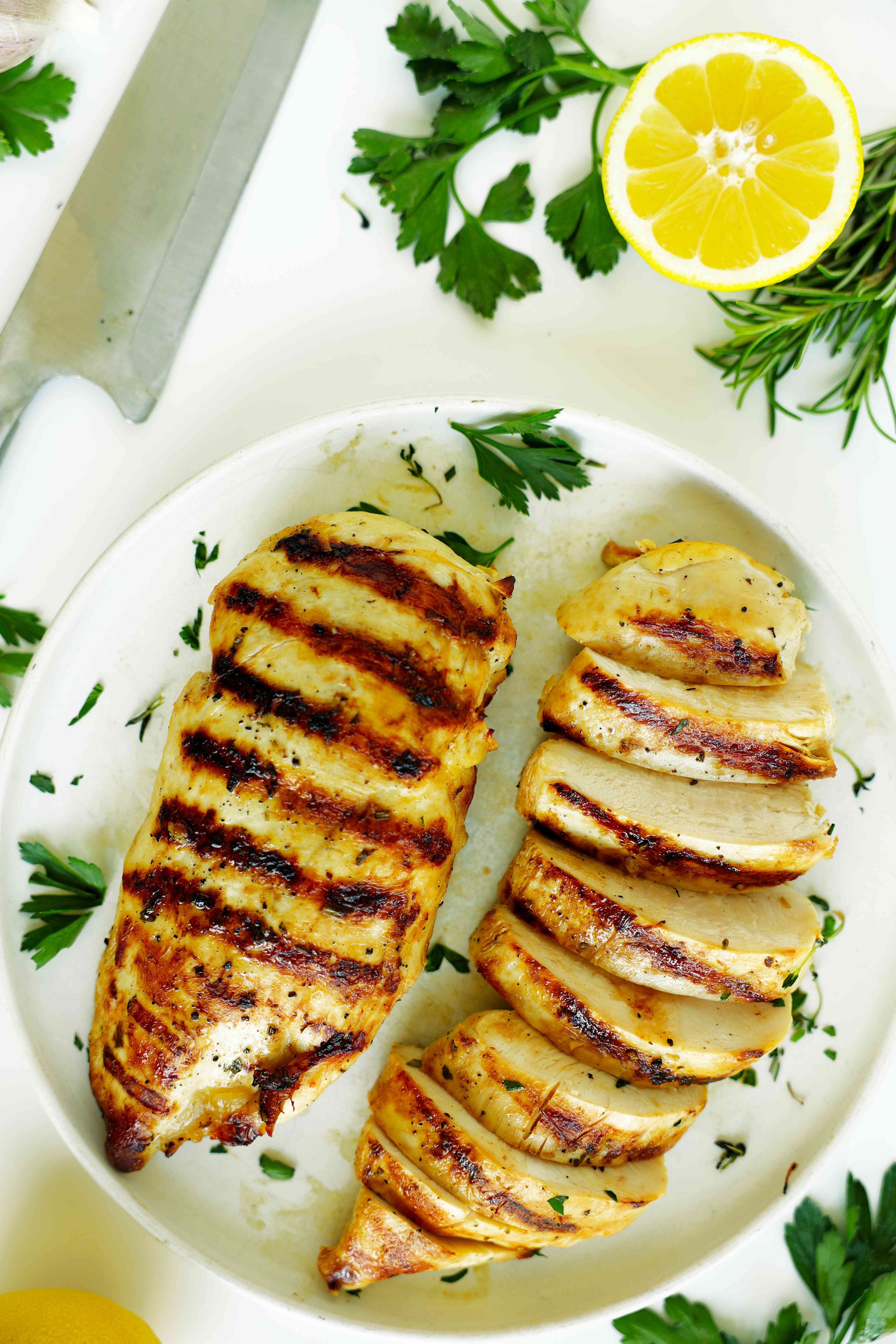 Juicy grilled chicken breast sliced and served with lemon and fresh herbs on a white plate
