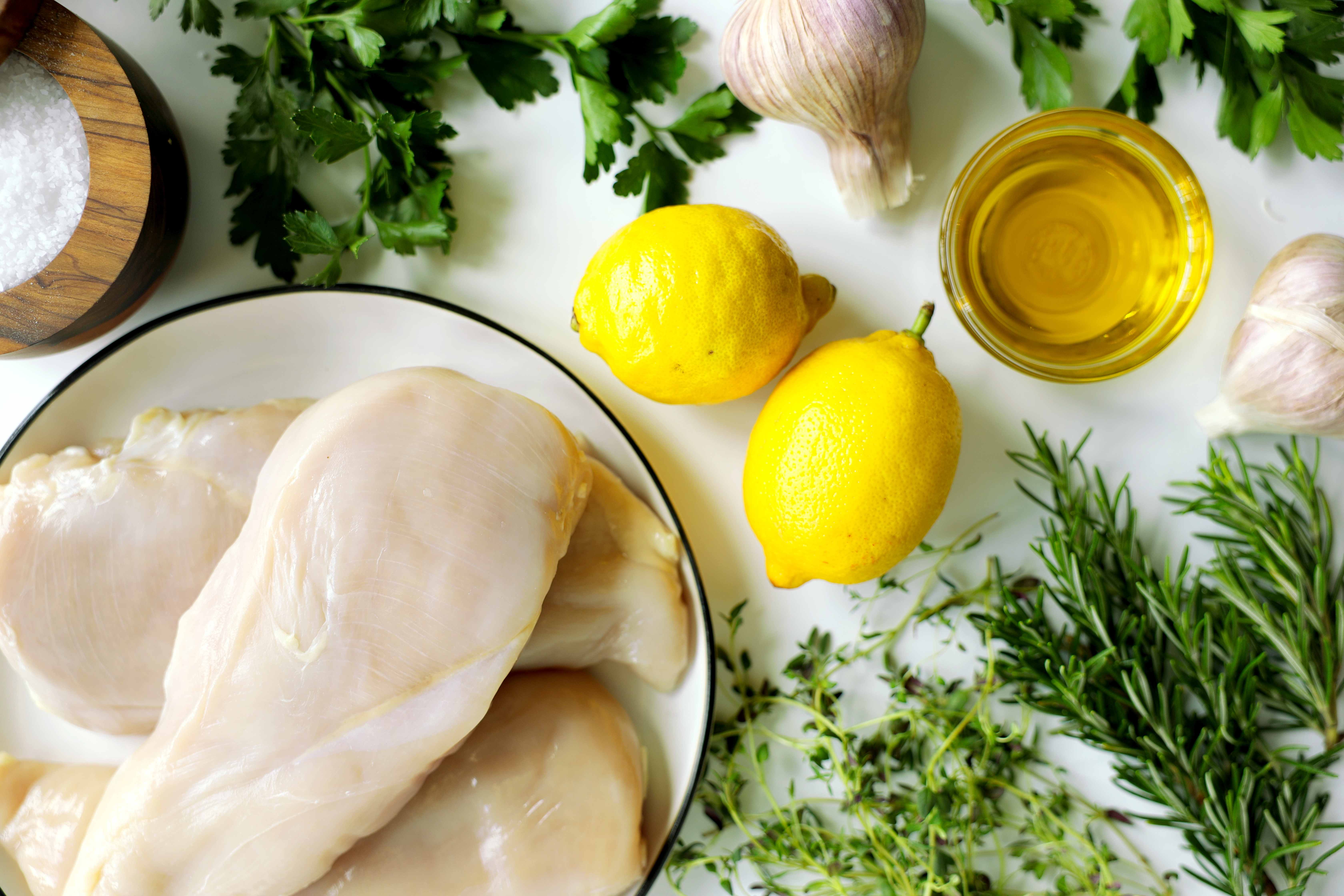 Herbs, lemon, garlic, oil, and salt laid out next to chicken breasts for the best chicken marinade recipe.