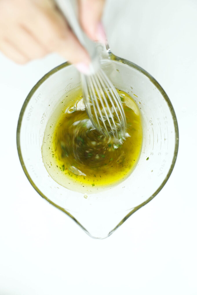 Whisking a garlic herb chicken marinade with olive oil, lemon juice, and herbs in a glass measuring cup