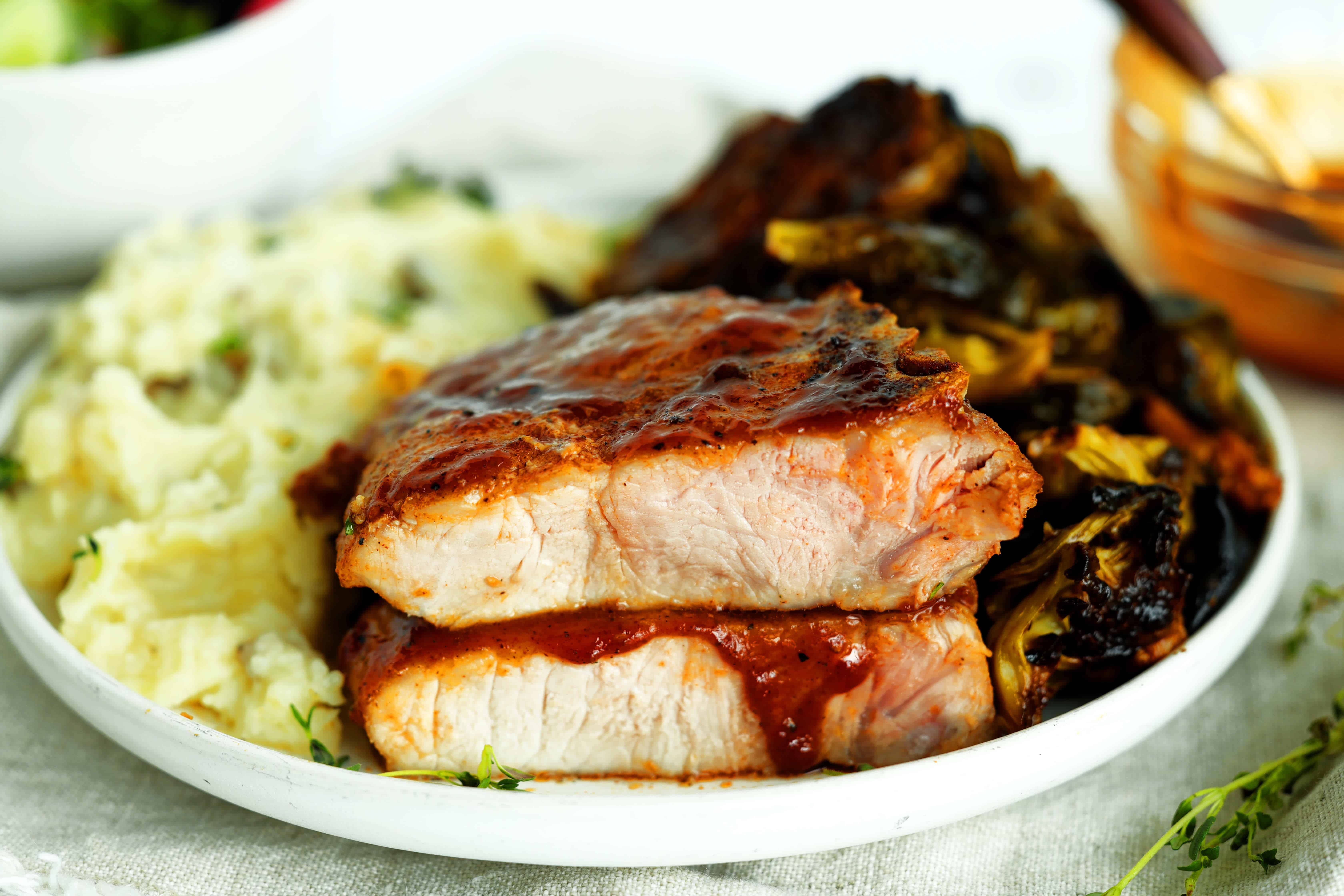 Sliced BBQ pork chop showing a juicy interior served with mashed potatoes and caramelized Brussels sprouts.