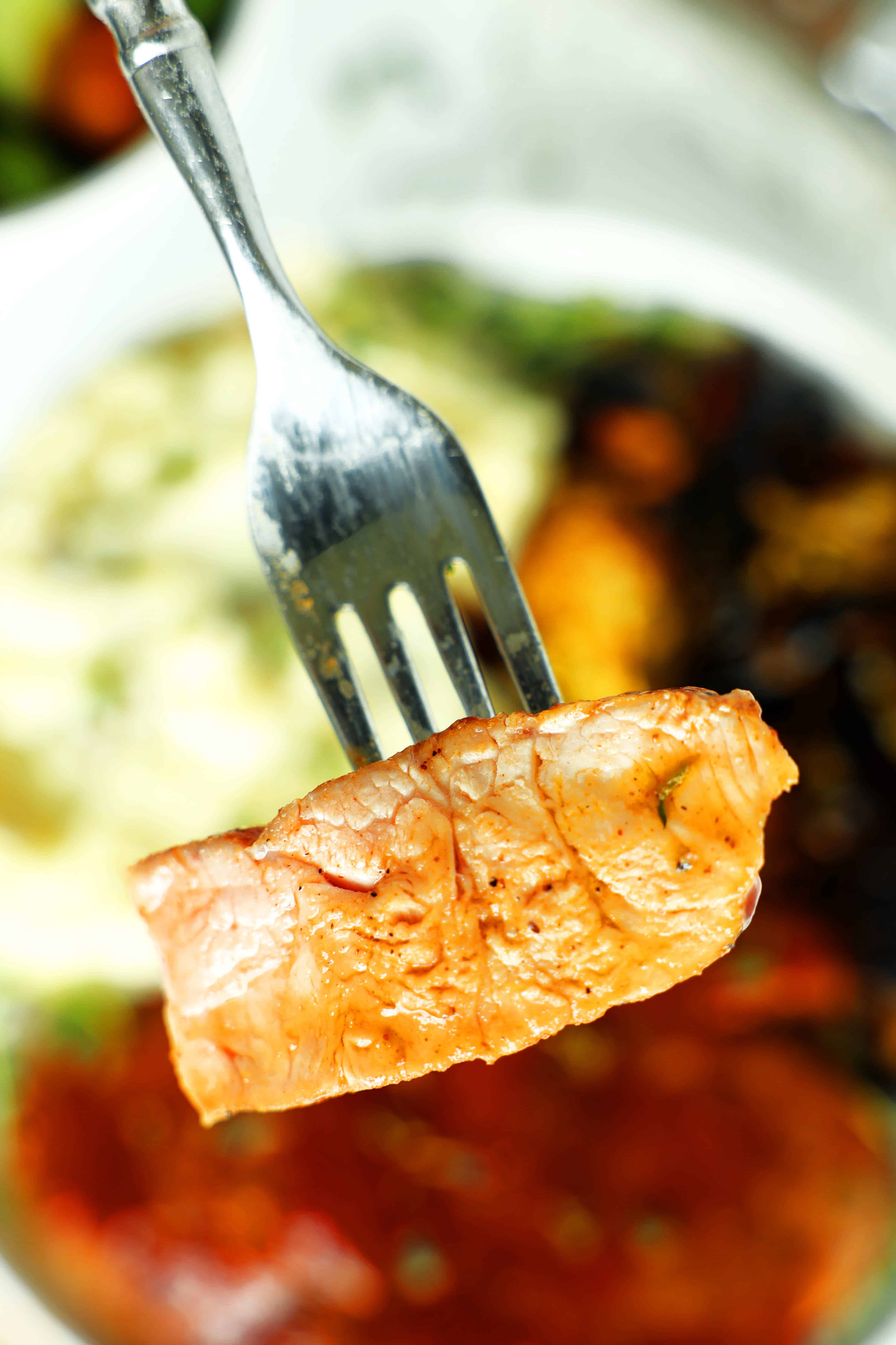 Juicy BBQ pork chop held on a fork with mashed potatoes and caramelized Brussels sprouts blurred in the background.