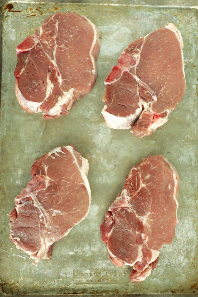 Four raw bone-in pork chops arranged on a rimmed baking sheet before seasoning.