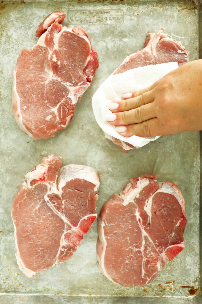 Paper towel being used to pat bone-in pork chops dry on a rimmed baking sheet.
