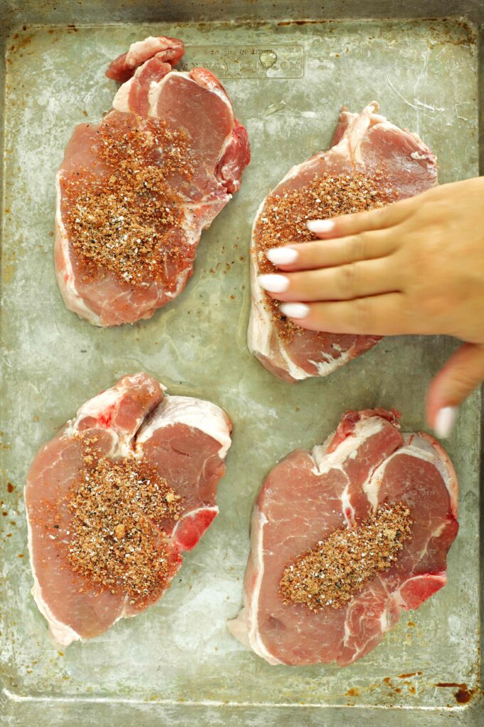 Hand sprinkling BBQ seasoning over raw bone-in pork chops on a baking sheet.