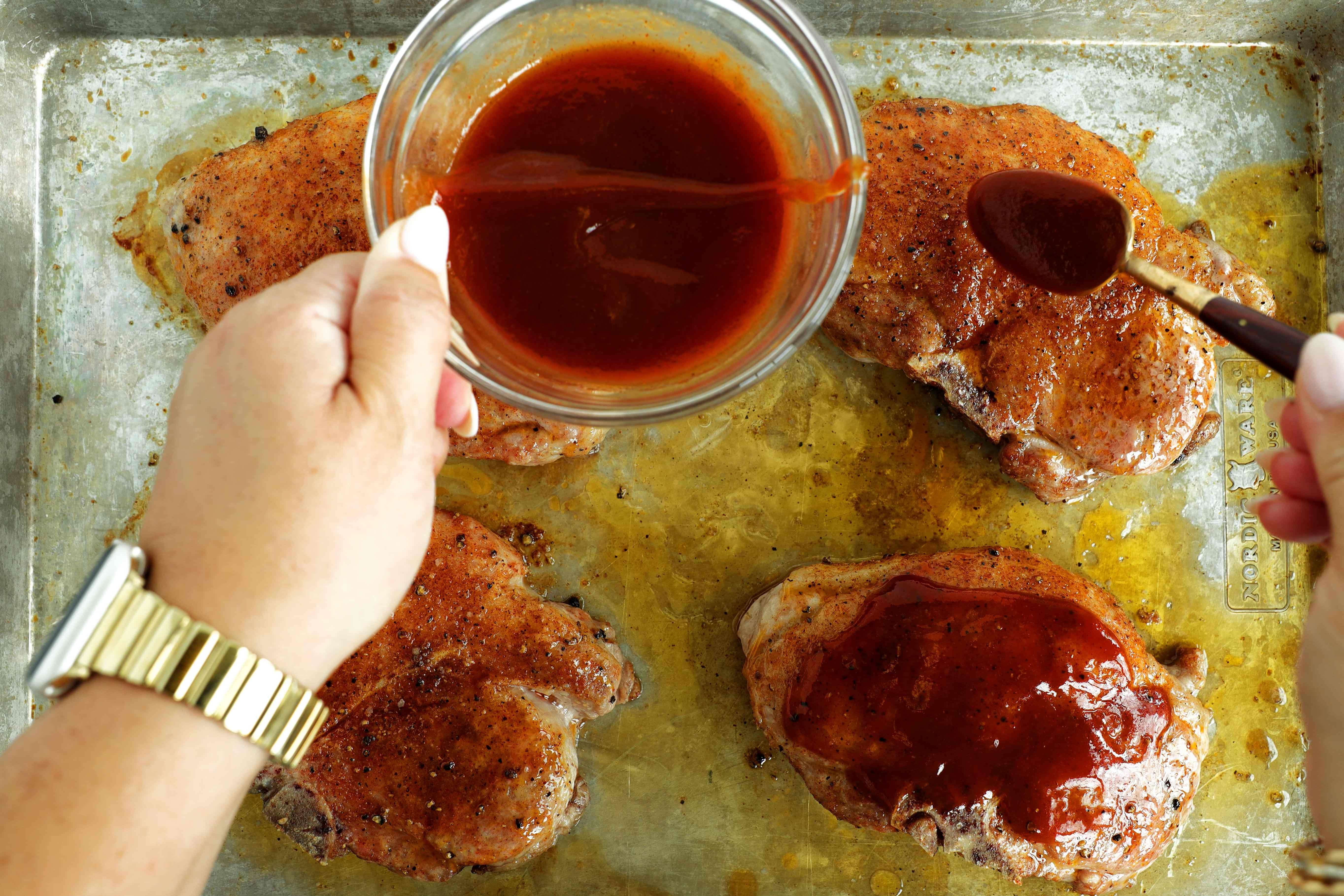 Hand spooning barbecue sauce over seasoned pork chops with a pastry brush.