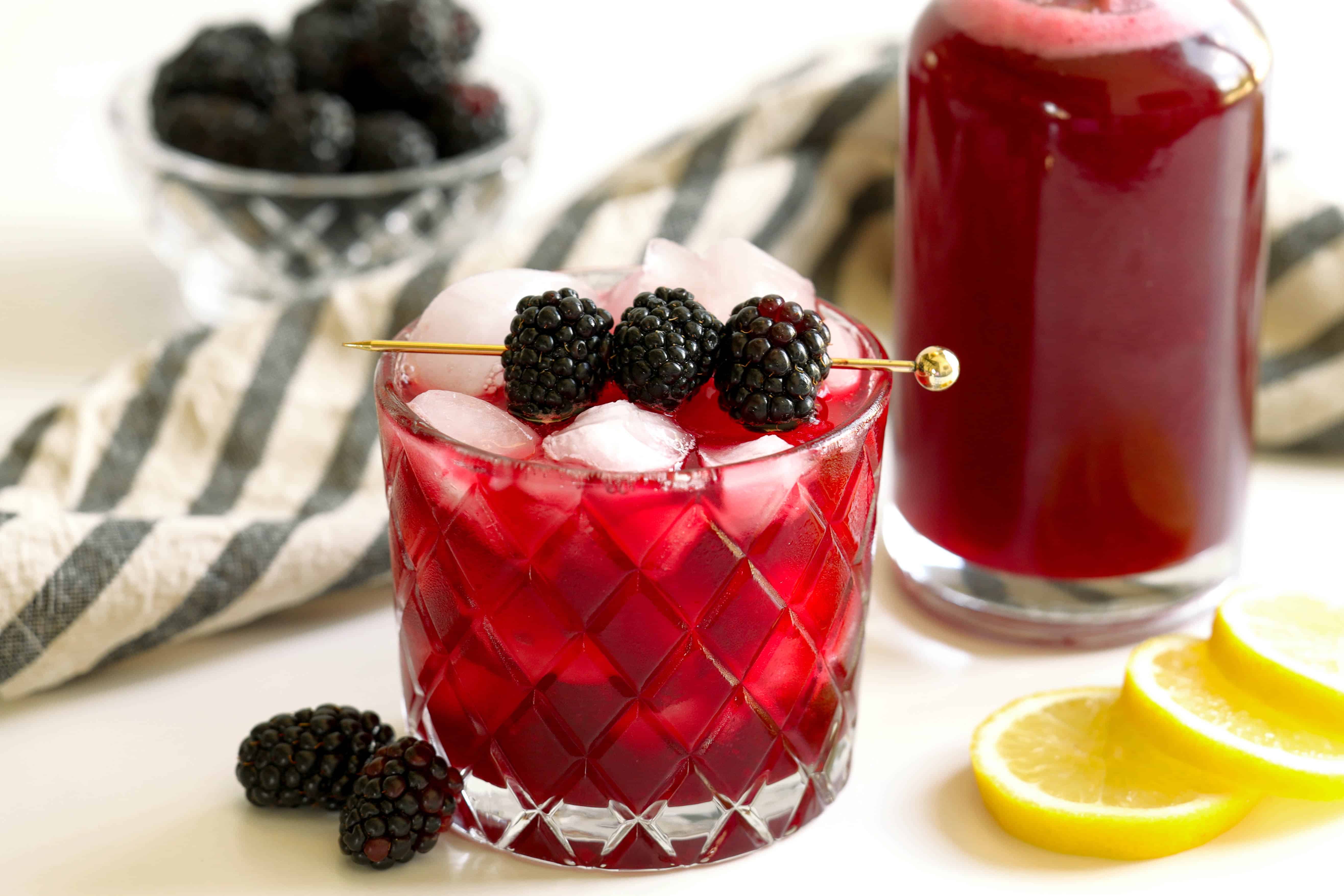 Glass of blackberry simple syrup cocktail over ice with fresh blackberries, lemon slices, and a bottle of homemade syrup
