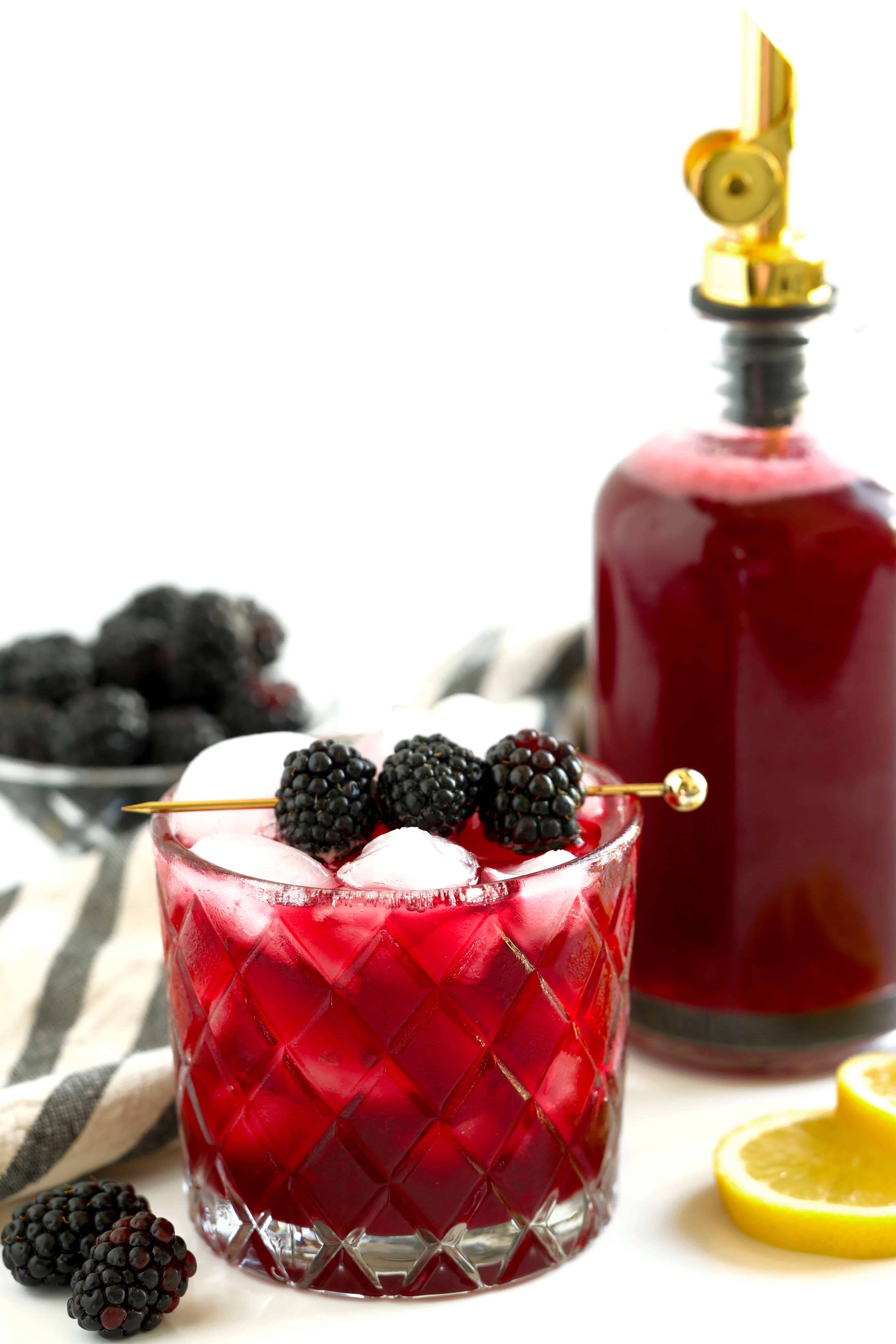 Glass of blackberry simple syrup cocktail over ice with fresh blackberries and a bottle of homemade syrup