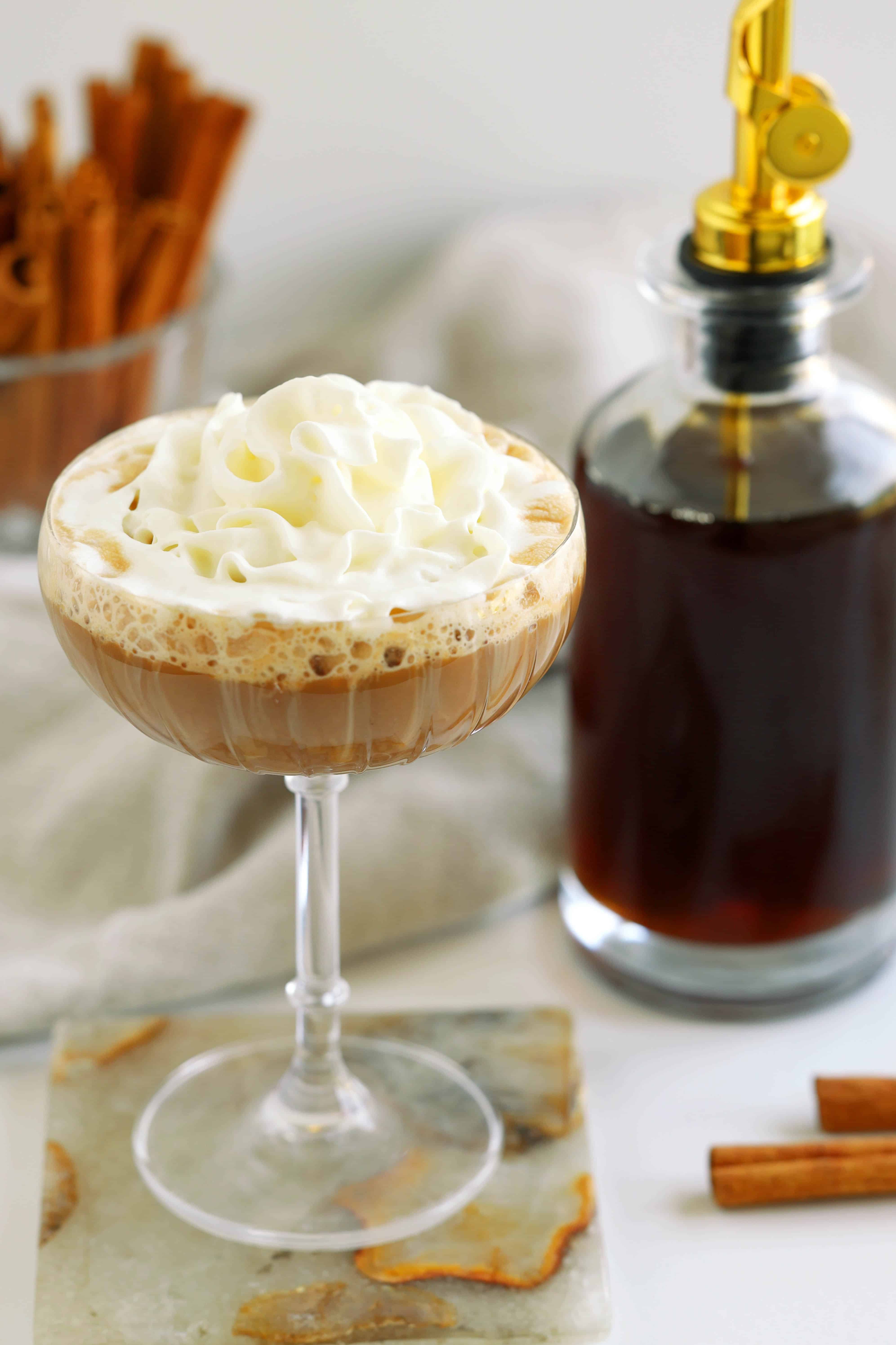 Cinnamon syrup beside a whipped cream topped coffee cocktail with cinnamon sticks in the background.