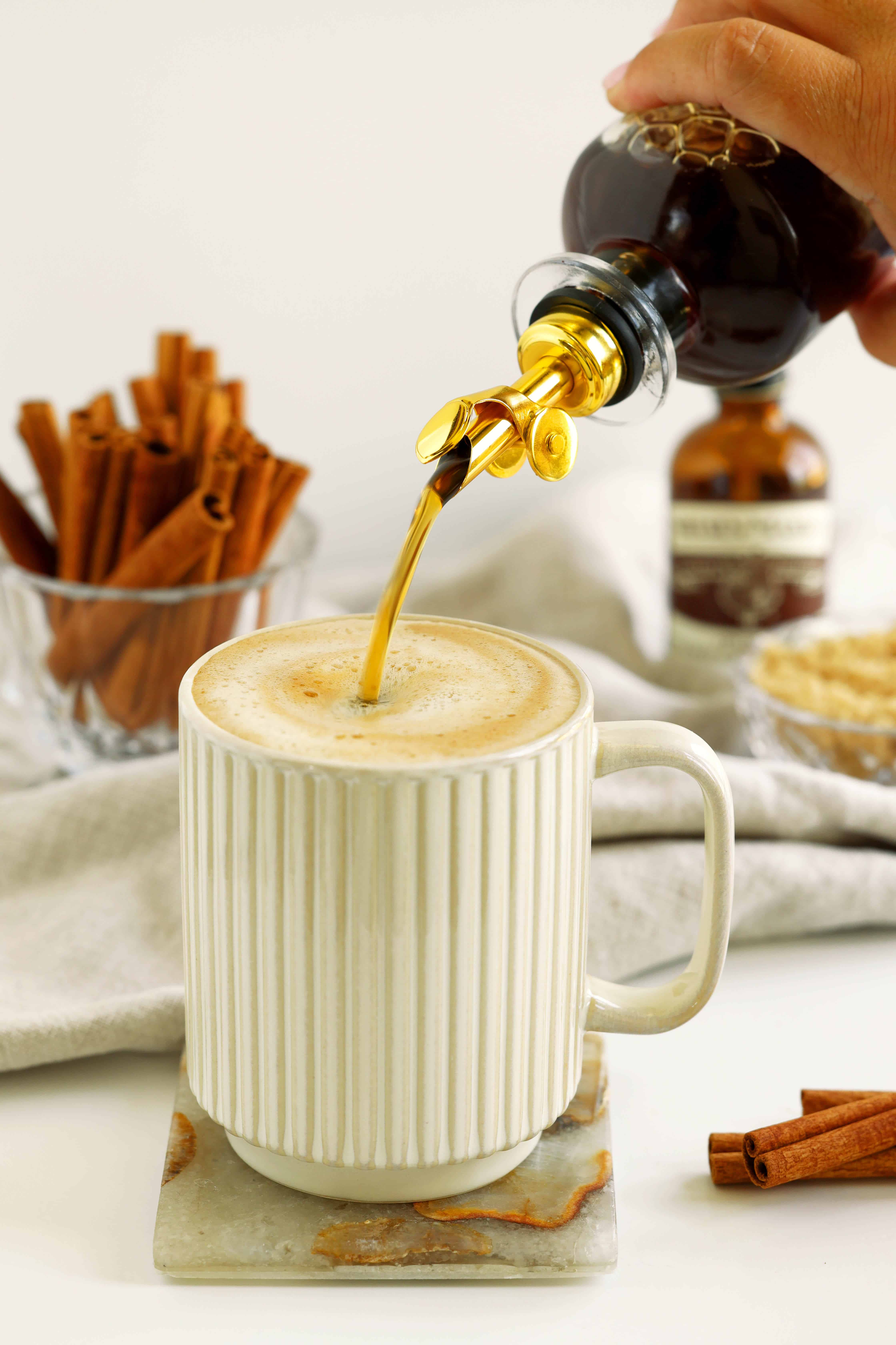 Cinnamon syrup being poured from a glass bottle into a foamy coffee with cinnamon sticks nearby.