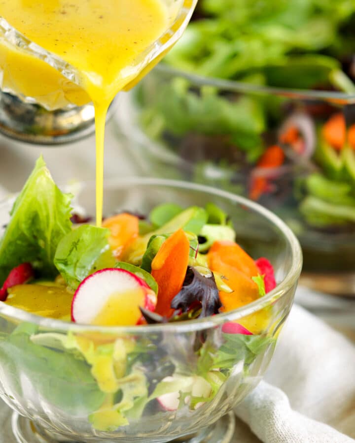 Dijon dressing being poured over a fresh green salad with radishes, carrots, and mixed greens