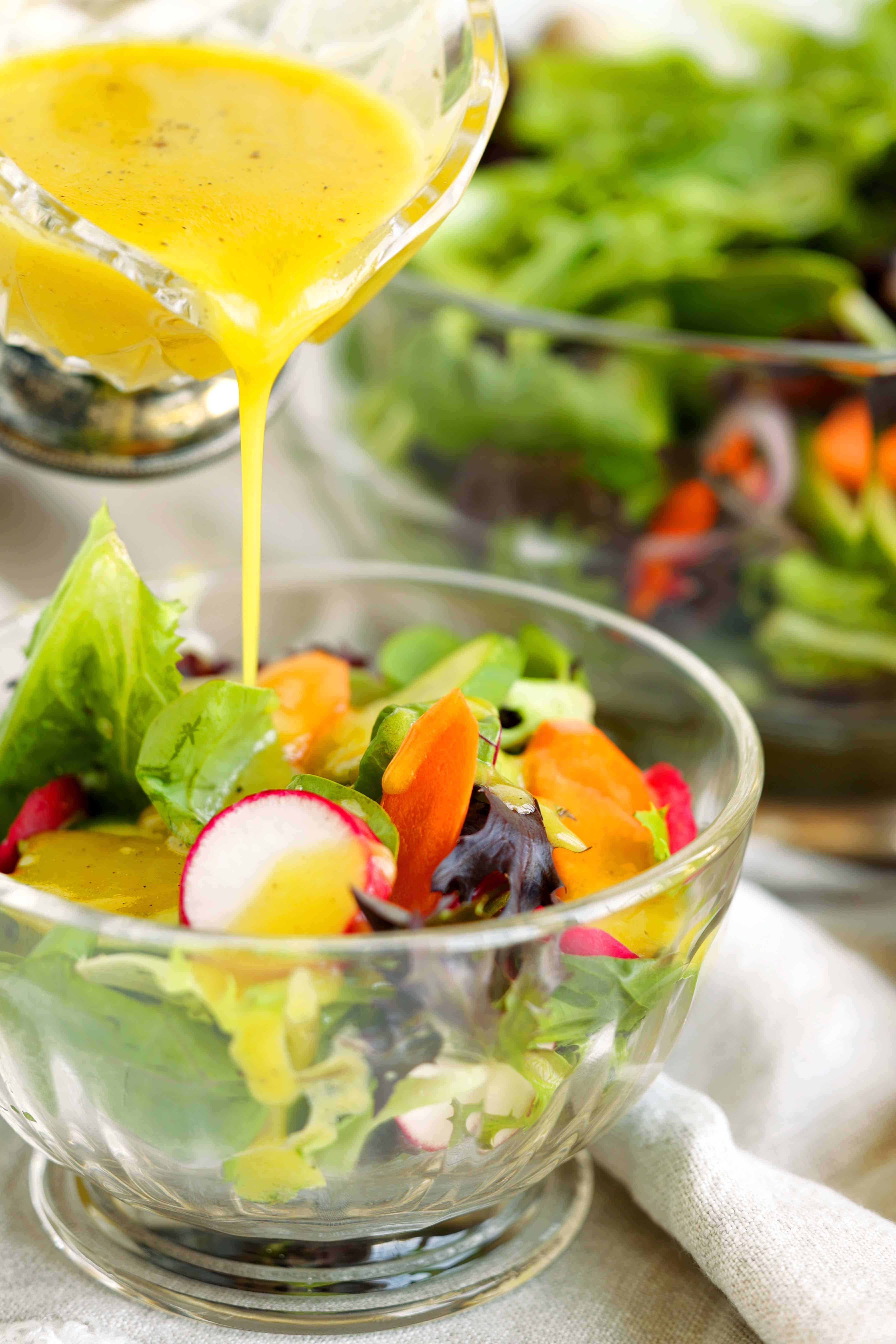 Dijon dressing being poured over a fresh green salad with radishes, carrots, and mixed greens