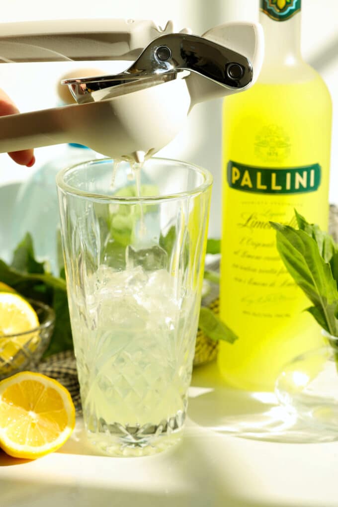 Fresh lemon juice being squeezed into a cocktail shaker with limoncello nearby