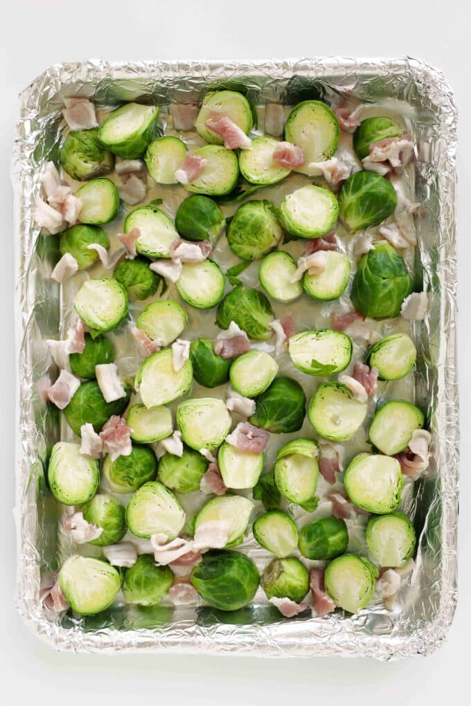 Brussels sprouts and chopped raw bacon spread across a foil-lined sheet pan