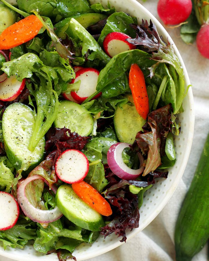Spring mix salad with cucumbers, radishes, carrots, and Dijon vinaigrette in a white serving bowl