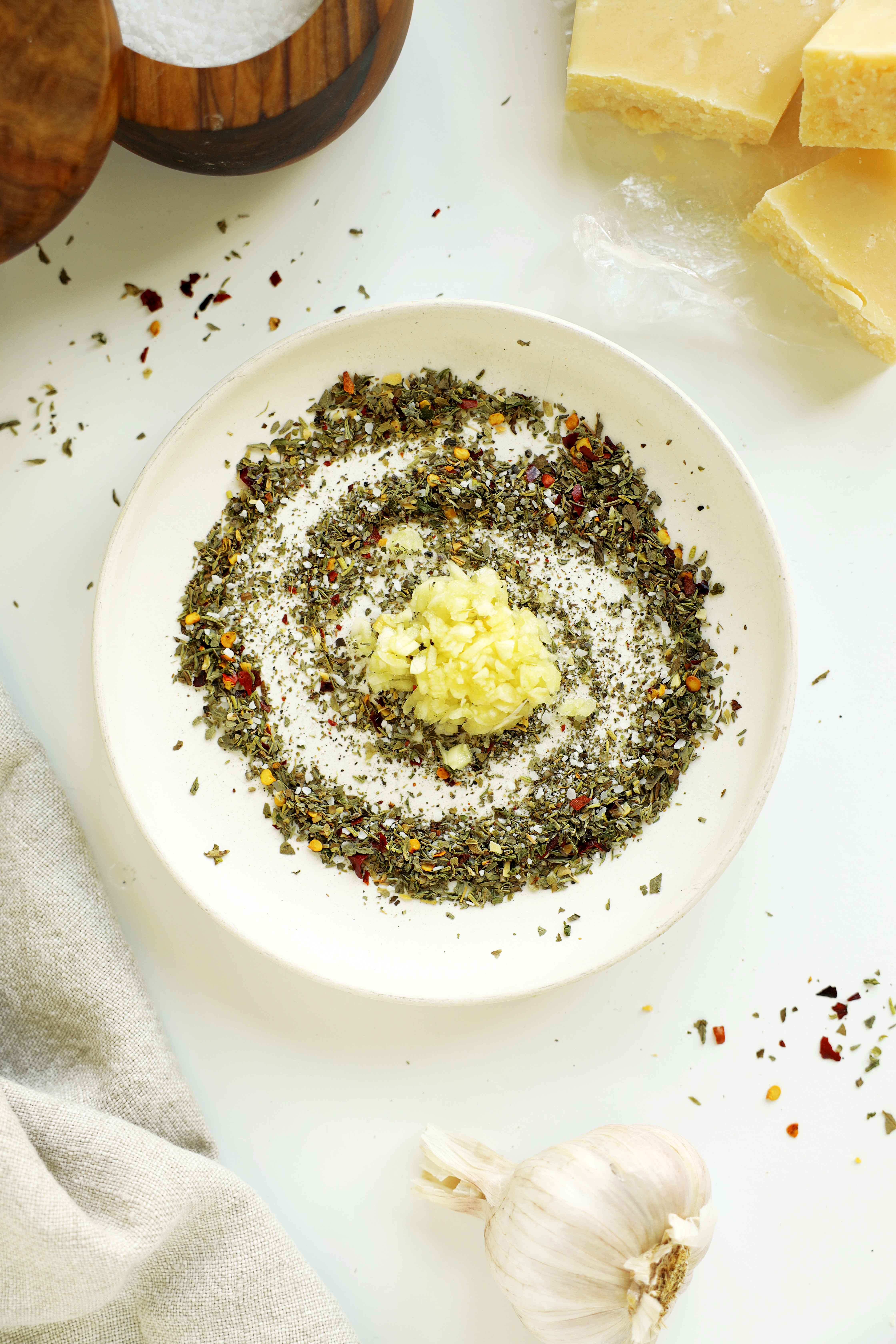 Minced fresh garlic added to a bowl of dried Italian herbs and seasonings.