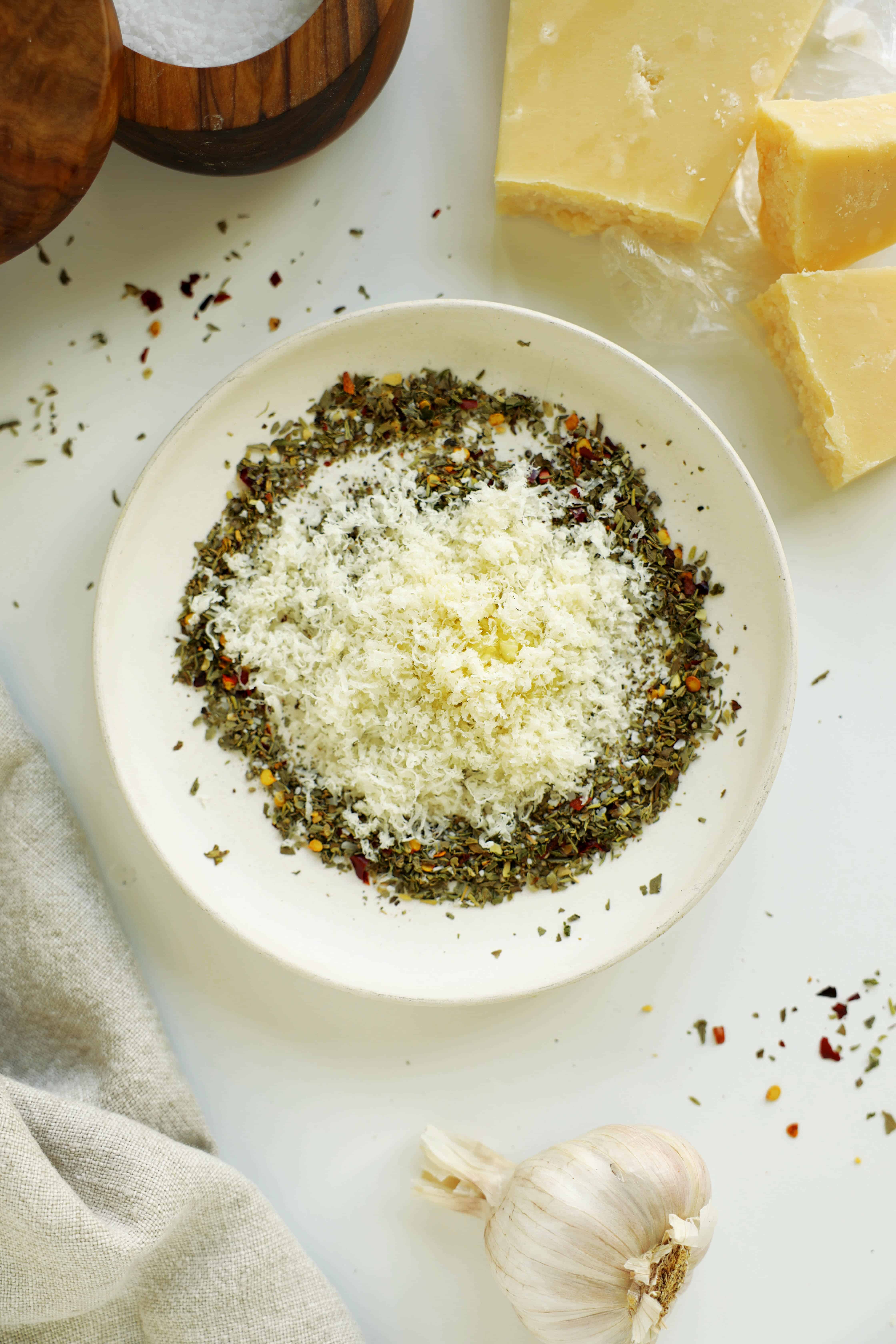 Grated Parmesan cheese added to dried Italian herbs and red pepper flakes in a white bowl.